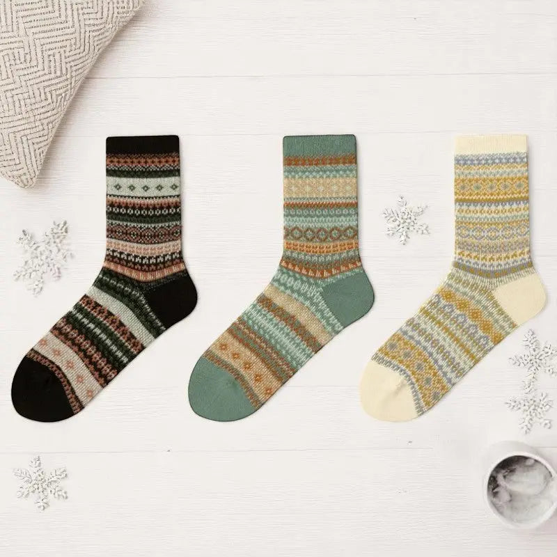 Trois paires de chaussettes d’hiver femme avec motifs nordiques : crème, vert sauge et noir sur fond bois clair.