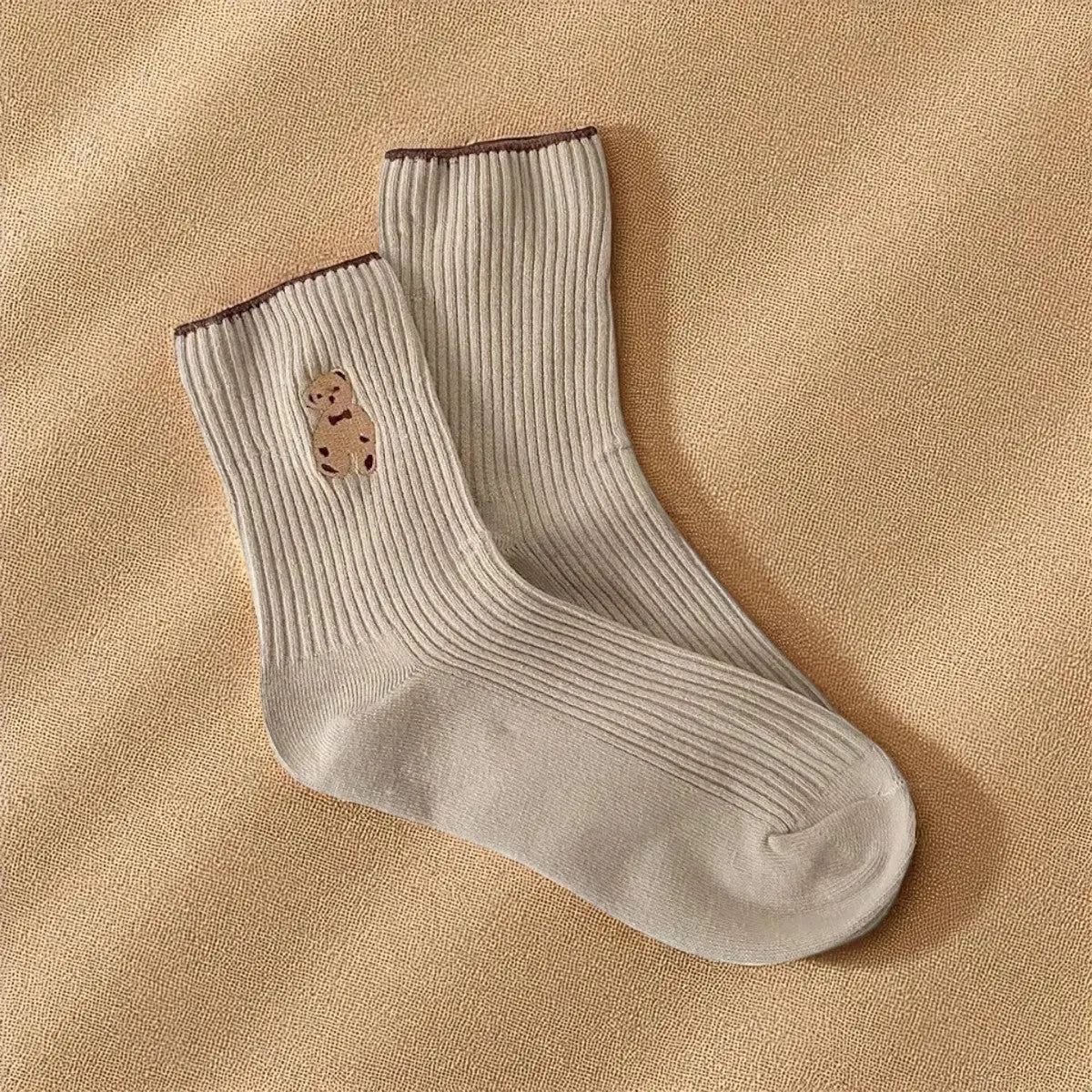 Chaussettes beiges côtelées avec motif ourson brodé, posées sur un tissu beige.