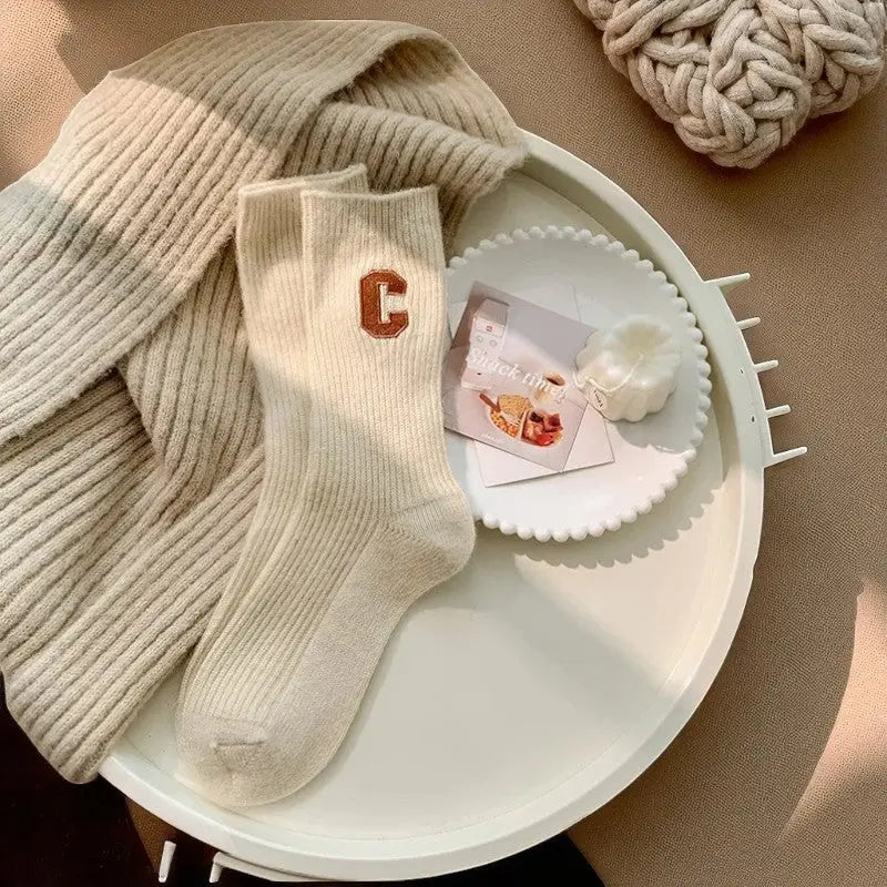 Chaussettes cachemire crème pour femme avec logo marron, posées sur un décor beige et texturé dans une ambiance chaleureuse.